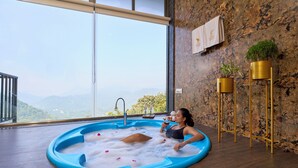 Una persona relajándose en una bañera de hidromasaje azul con pétalos de rosa, vista a las montañas y una pared rústica con detalles dorados.
