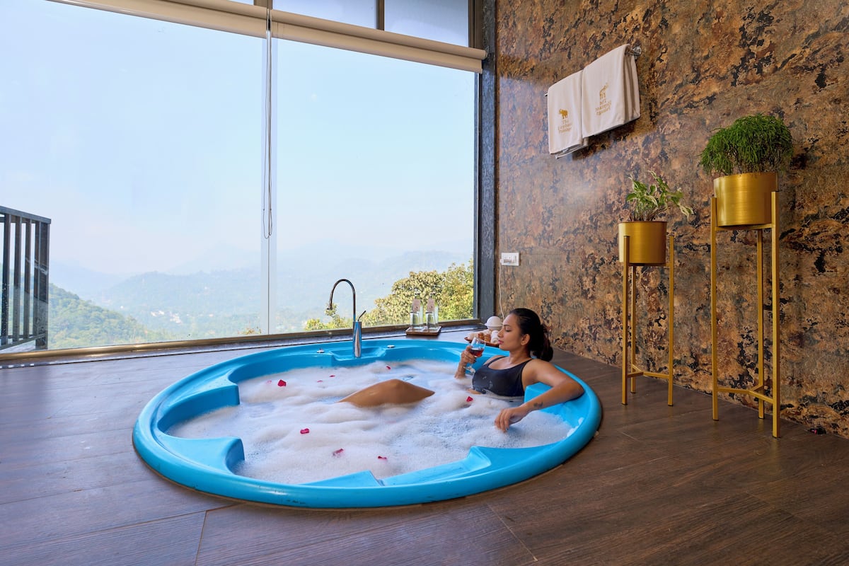 A person relaxing in a blue hot tub with rose petals, a view of mountains, and a rustic wall with gold decor.