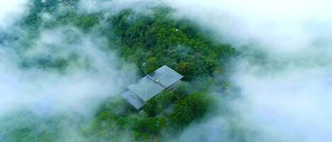 Vista aérea de un edificio de una planta rodeado de bosque denso, parcialmente cubierto por nubes.