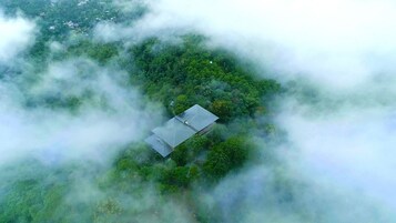 An aerial view of a single-story building surrounded by dense forest, partially obscured by clouds.