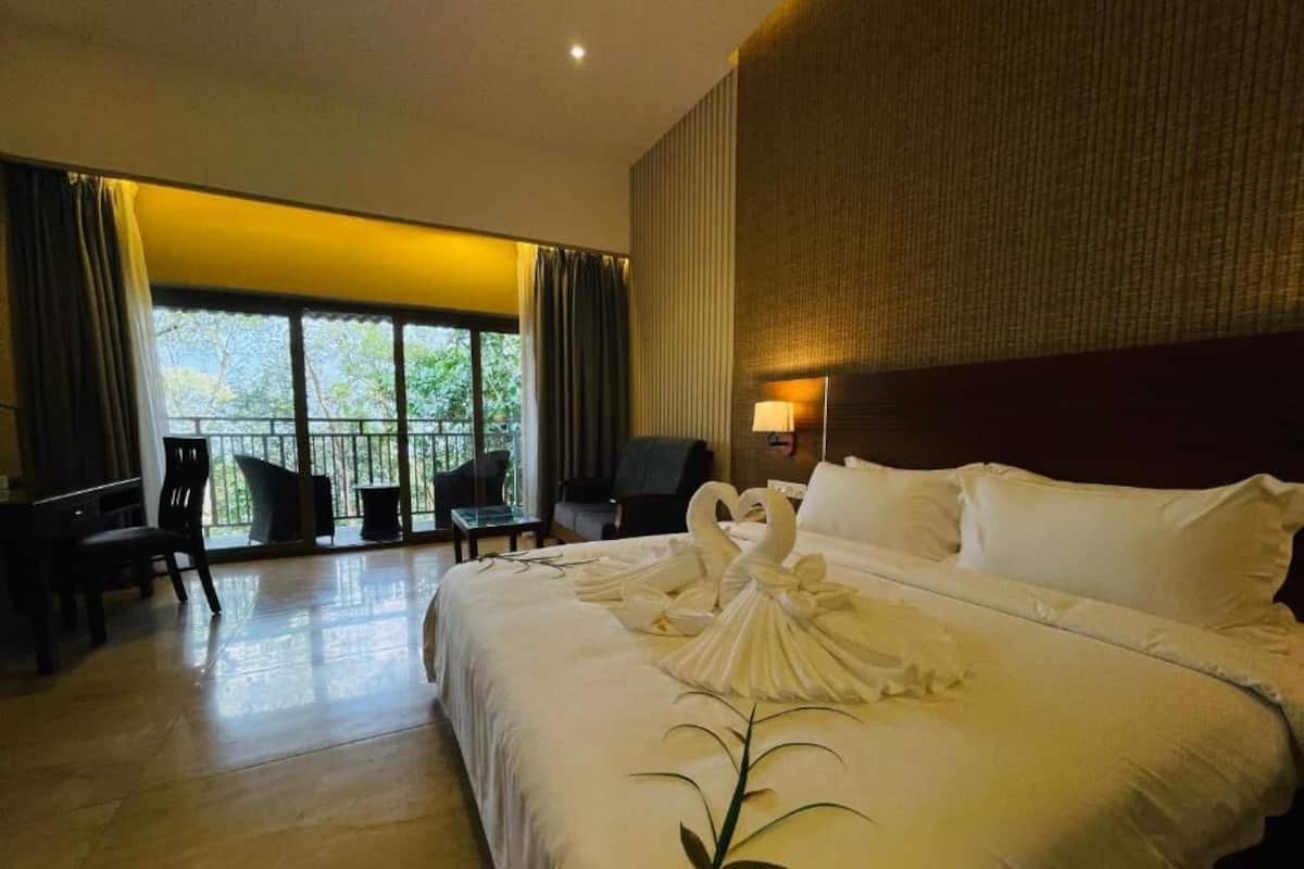 A hotel room with a large bed, a desk, and a chair. There is a balcony with a view of trees.