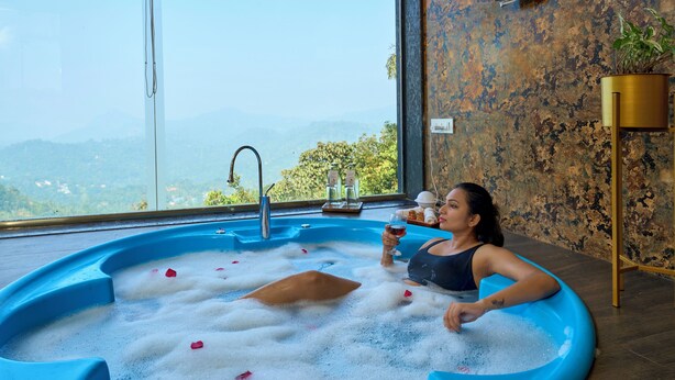 Una persona relajándose en un jacuzzi azul con pétalos de rosa, vista a las montañas y una pared rústica con una planta.