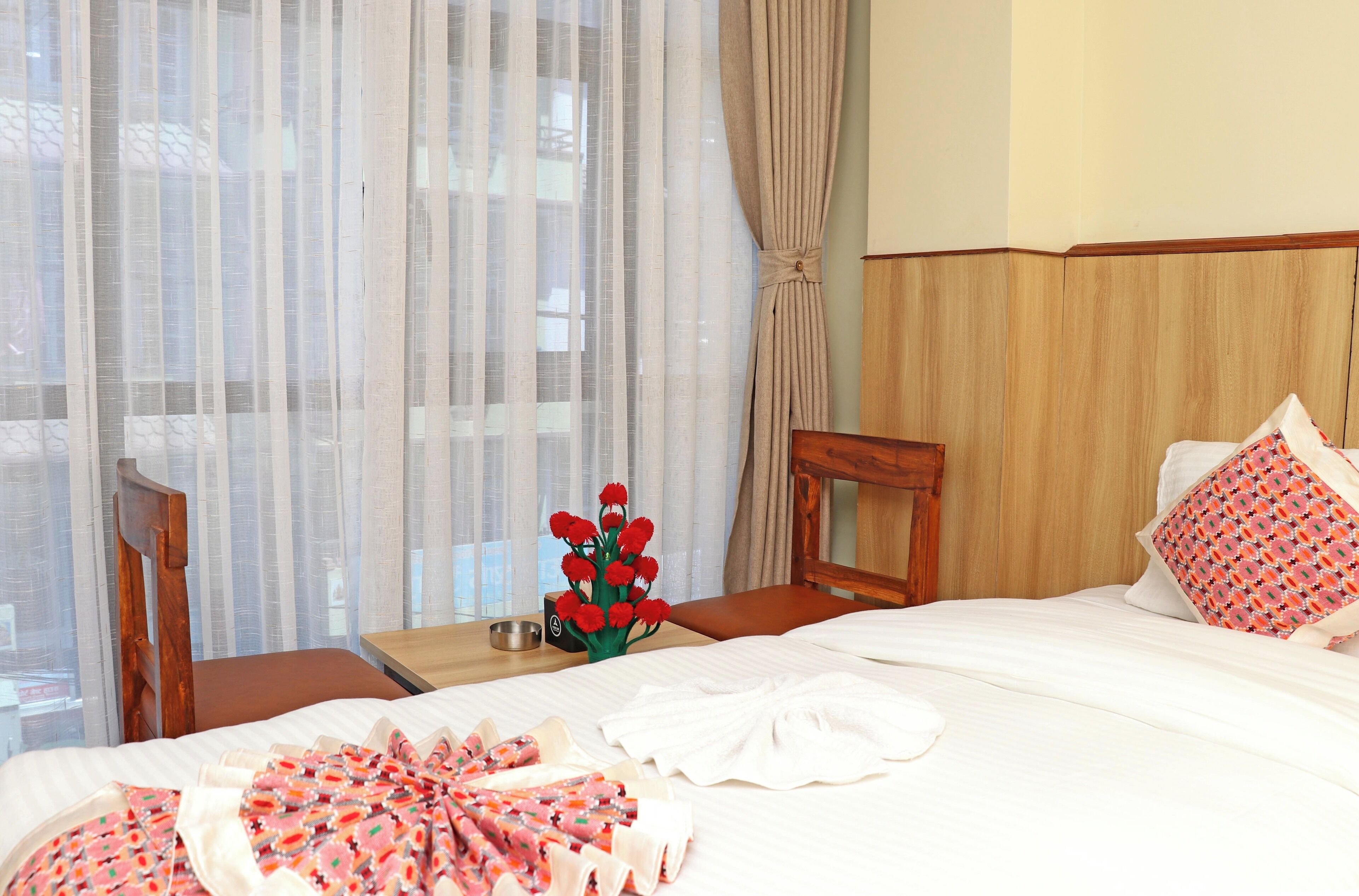 A hotel room with a bed, a wooden nightstand, a chair, a vase with red flowers, and a window with sheer curtains.