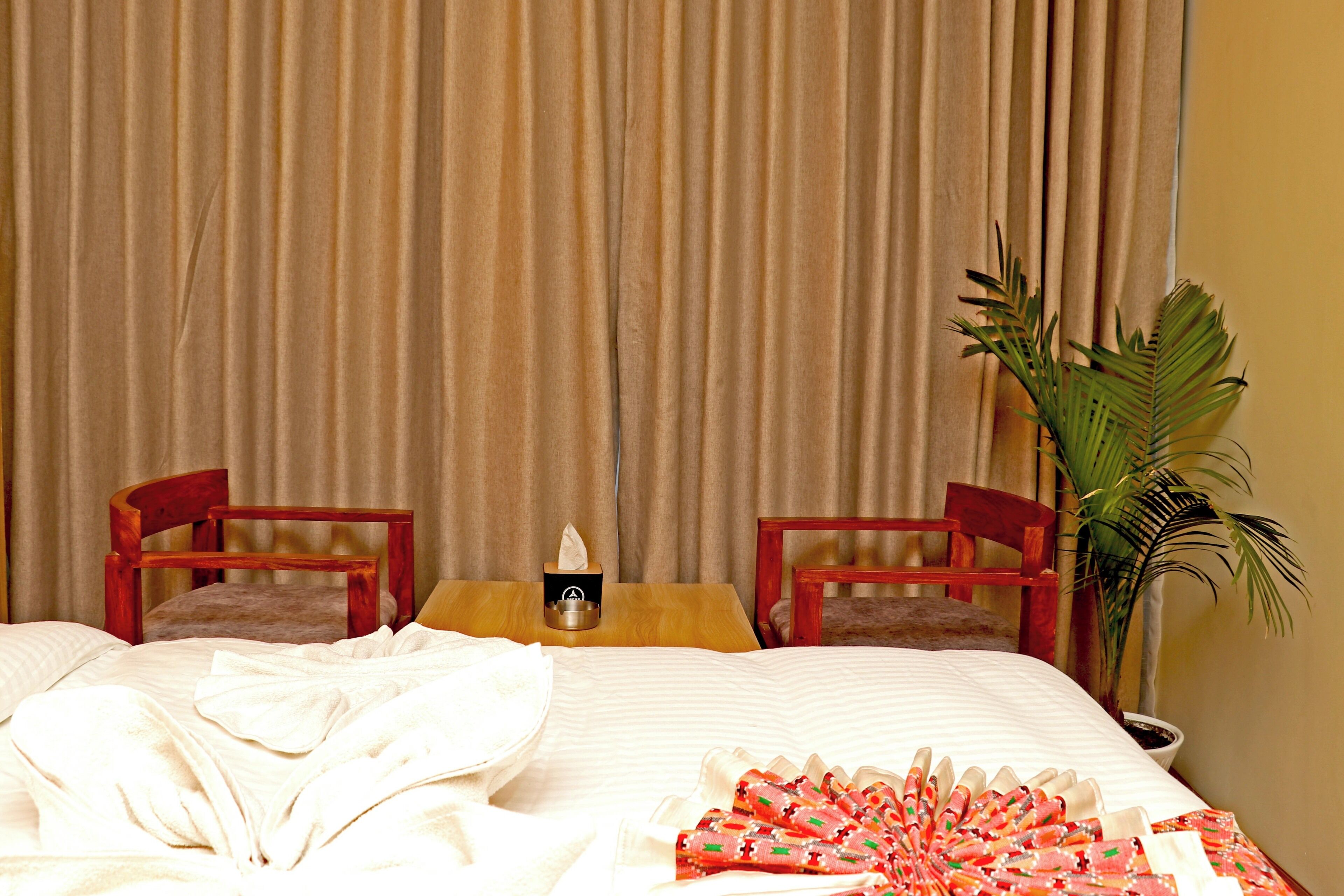 Een hotelkamer met een bed, twee houten stoelen, een klein tafeltje met zakdoosjes en een kamerplant.