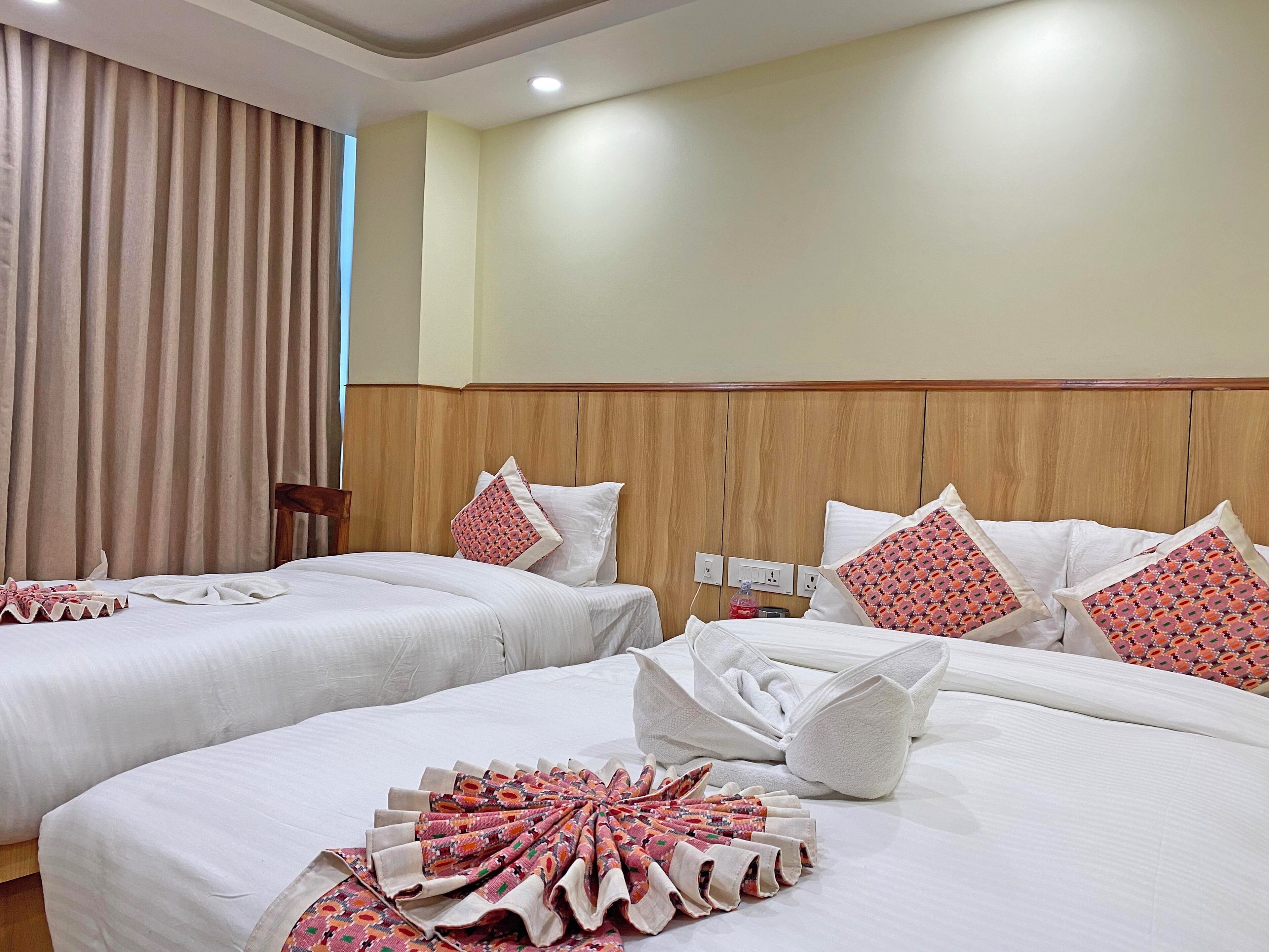 A hotel room with two beds, beige curtains, wooden headboard, white bedding, and patterned pillows.