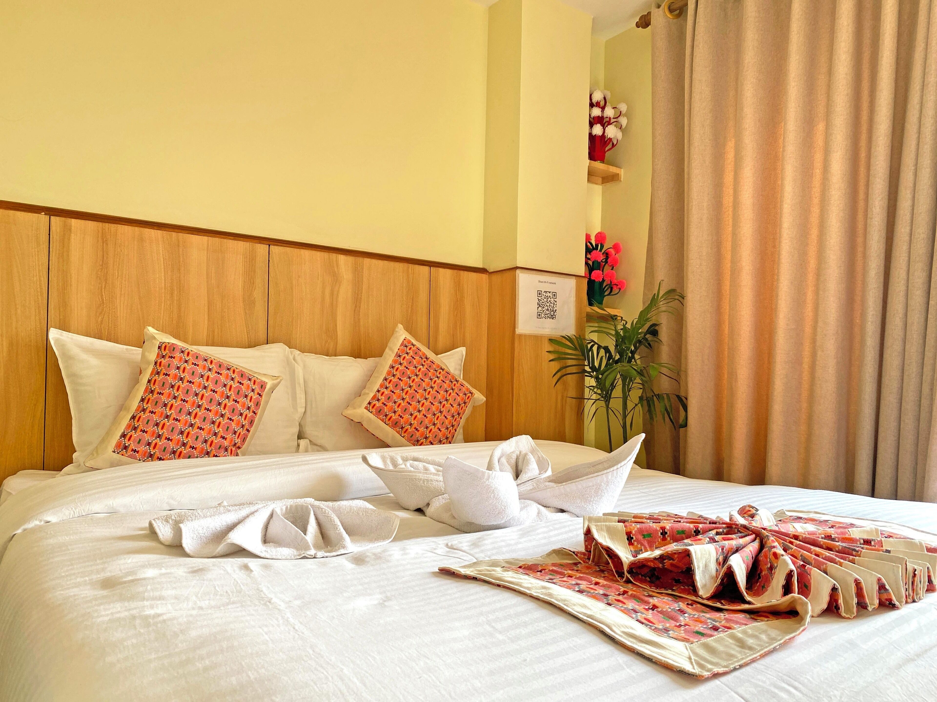 Una cama bien hecha con sábanas blancas, almohadones decorativos, un cabecero de madera y una planta en la esquina.