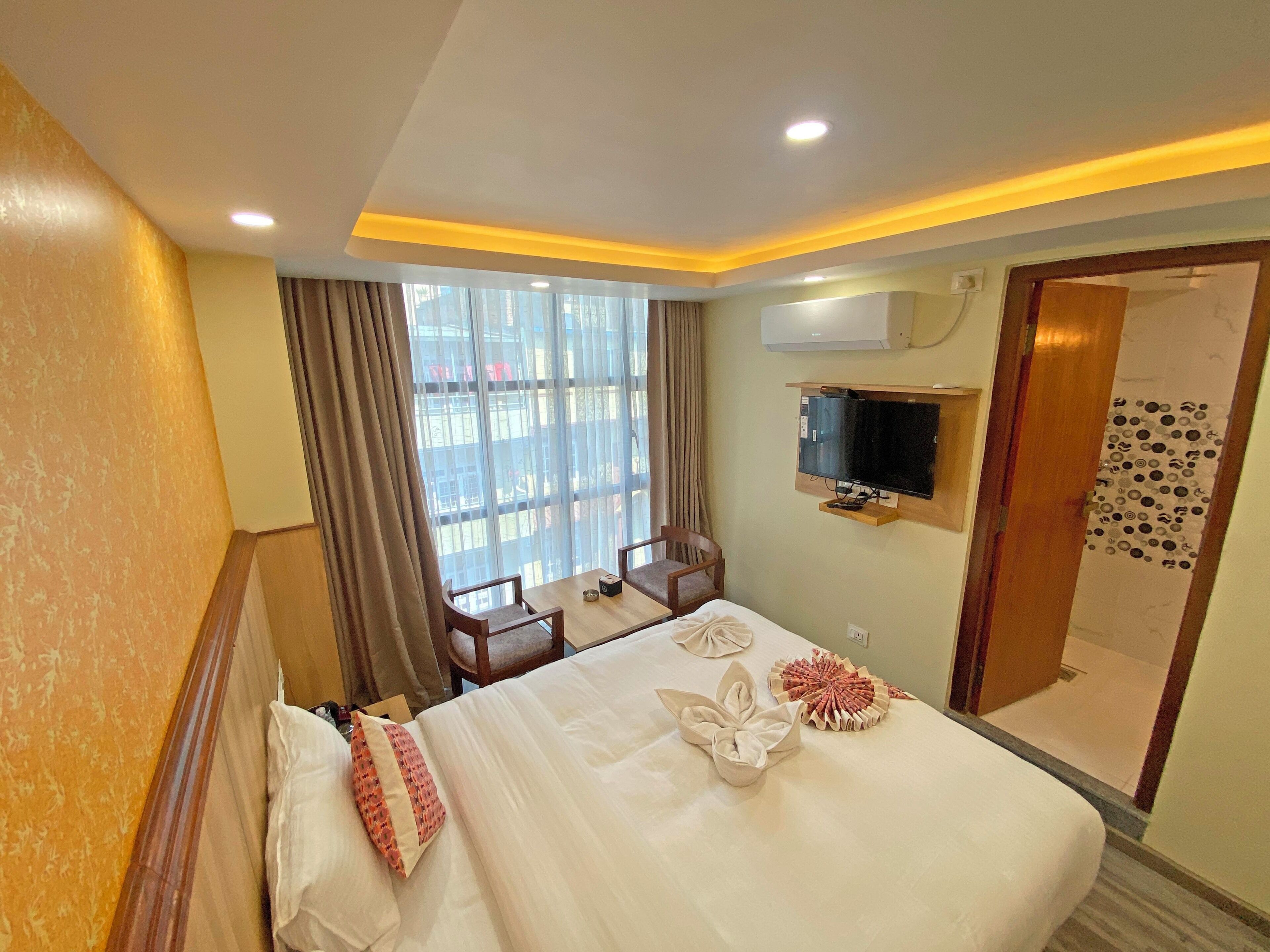 Una habitación de hotel con cama, televisor montado en la pared, ventana con cortinas y zona de ducha.
