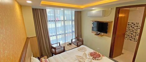 Una habitación de hotel con cama, televisor montado en la pared, ventana con cortinas y zona de ducha.