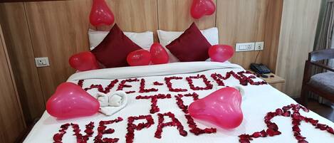 A bed with a welcome message and heart-shaped balloons, a telephone on a bedside table, and a wooden headboard.