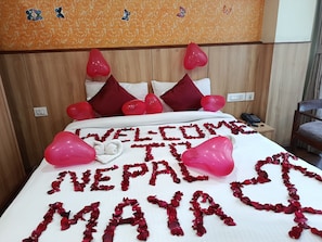 Una cama con un mensaje de bienvenida y globos con forma de corazón, un teléfono en la mesita de noche y un cabecero de madera.