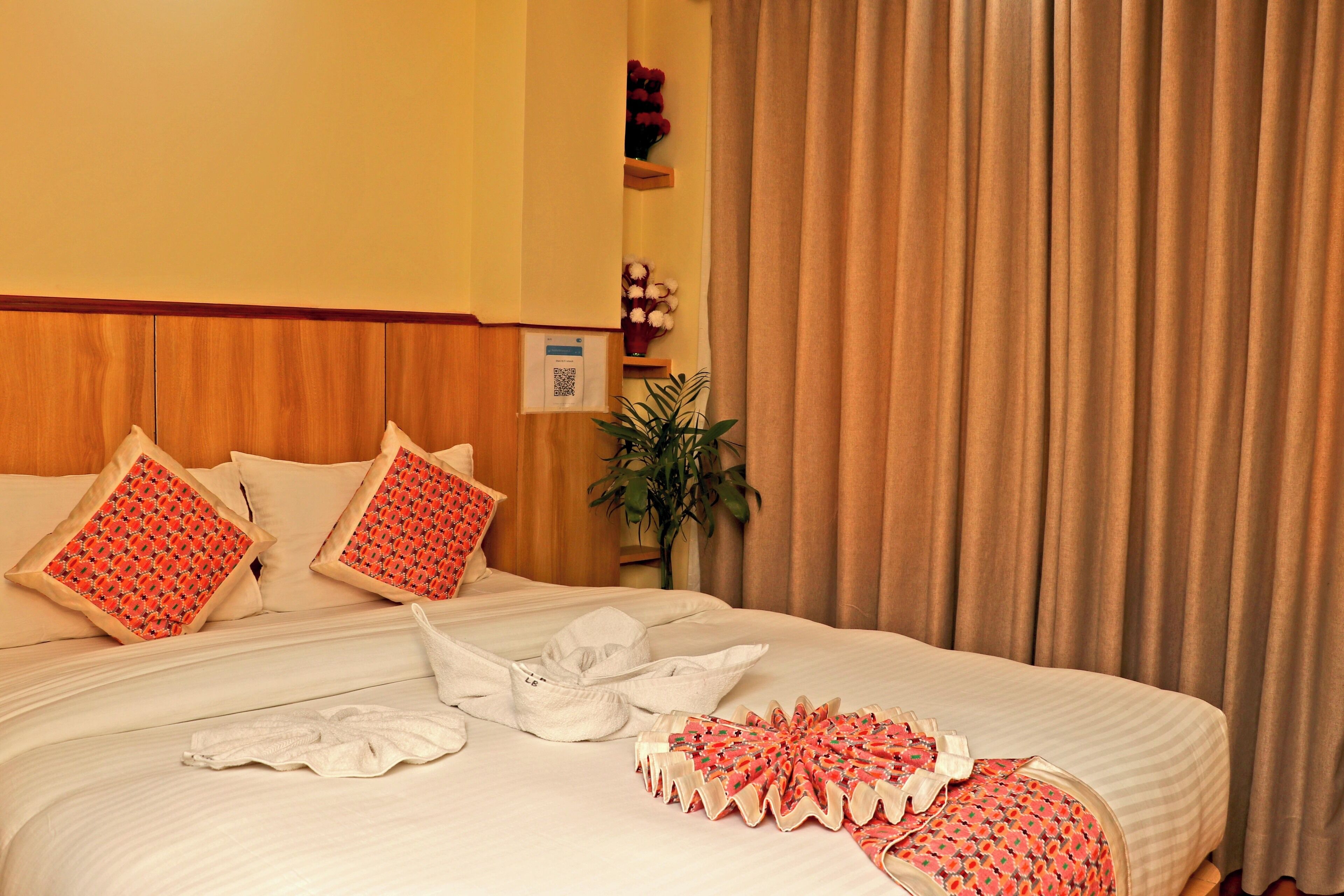 A neatly made bed with white linens and red patterned pillows, a bedside plant, and a wooden headboard.