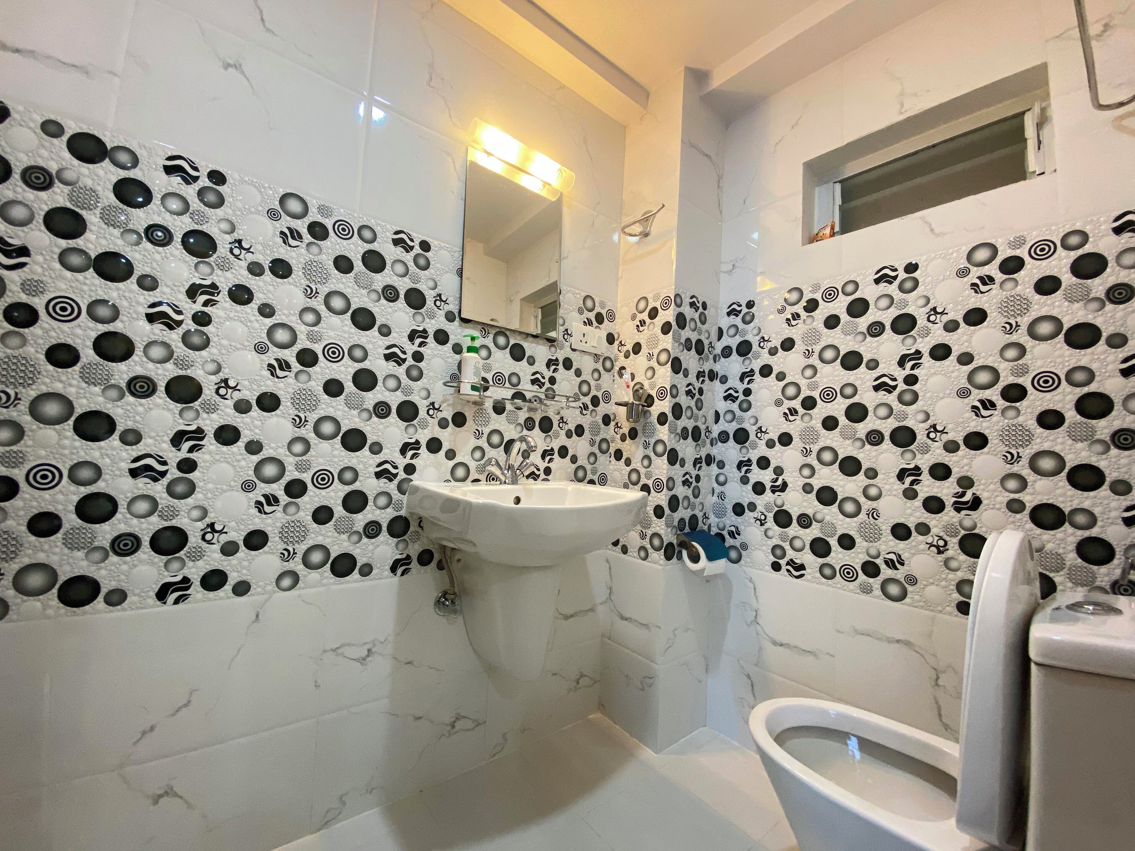 Un baño con lavamanos blanco, inodoro y espejo sobre el lavamanos. Las paredes están cubiertas con un diseño de azulejos a blanco y negro.