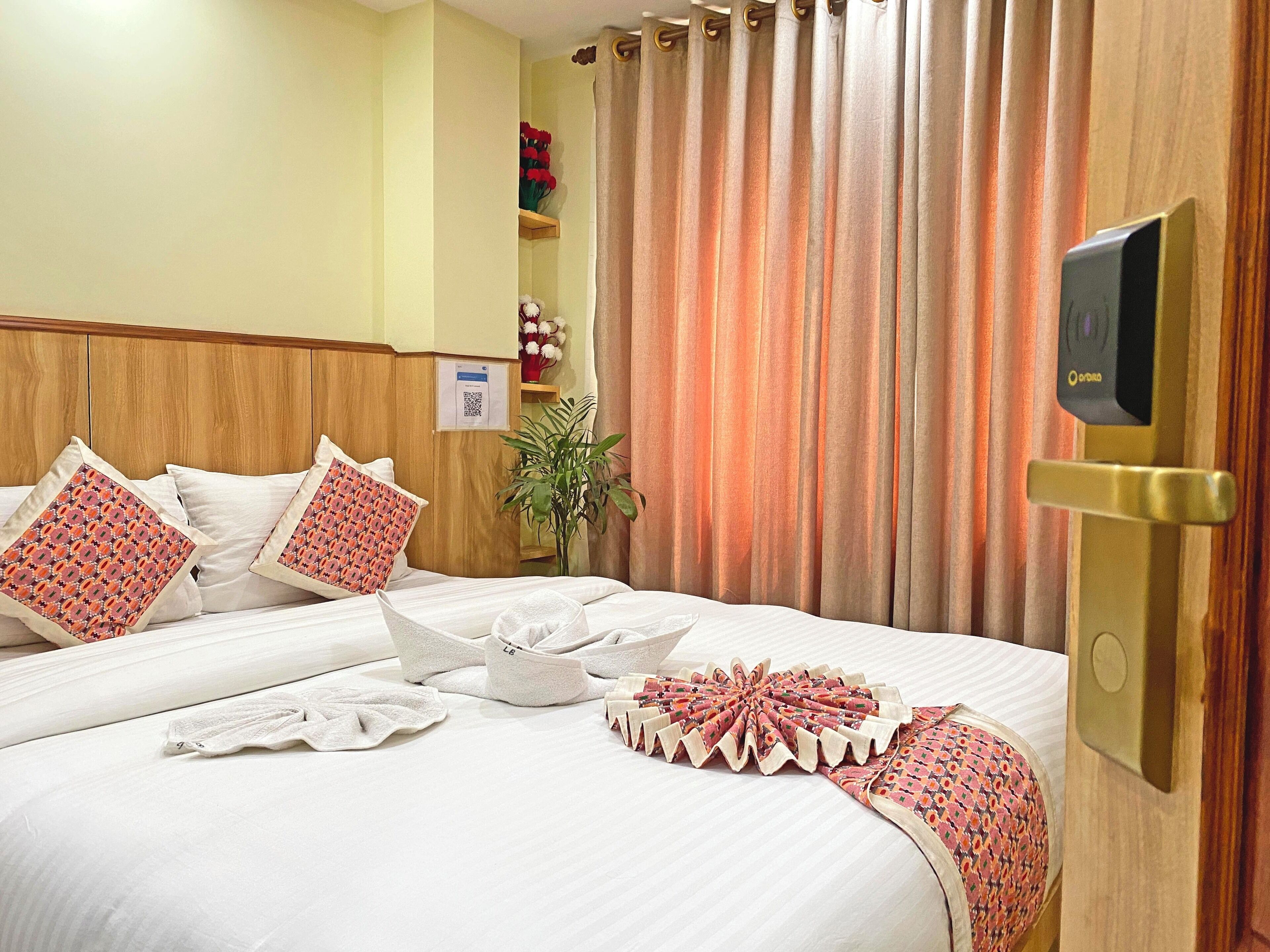 A hotel room with a bed, pillows, a plant, a TV, and a door with a lock.