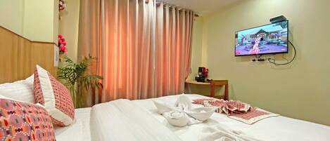 Habitación de hotel con una cama bien hecha, un televisor en la pared y una pequeña mesa con un teléfono y un objeto decorativo.