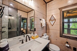 A modern bathroom with a large mirror, a sink with a towel holder, a toilet, and a window with blinds.
