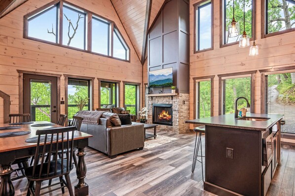 A spacious living area with a fireplace, wooden floors, and large windows offering views of the outdoors.