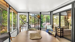Un espace repas moderne avec une table ronde en bois, des chaises et un set de table en fibres tressées. De grandes fenêtres offrent une vue sur les arbres et une terrasse.