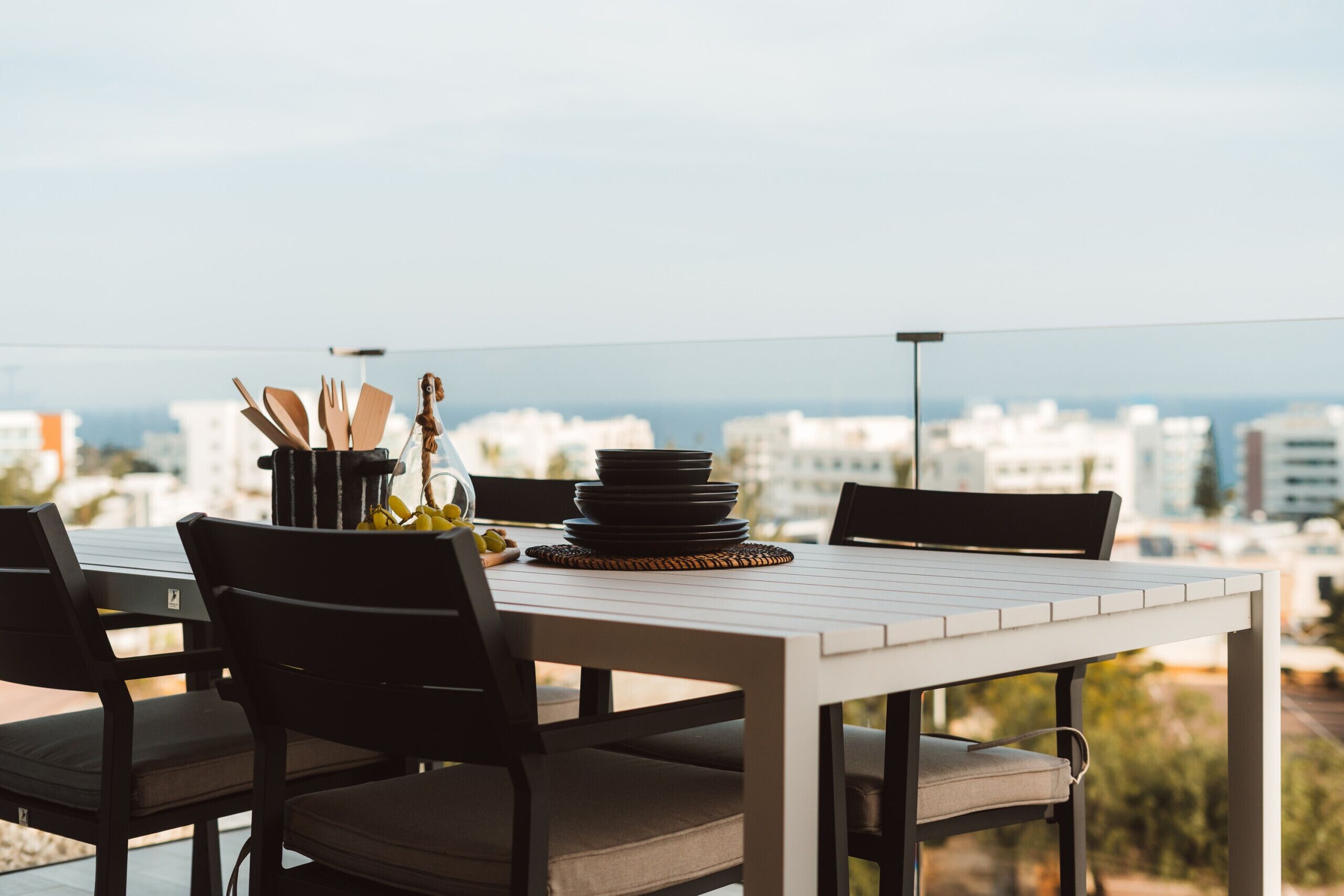 A dining table with chairs and a stack of plates, set on a balcony with a city view.