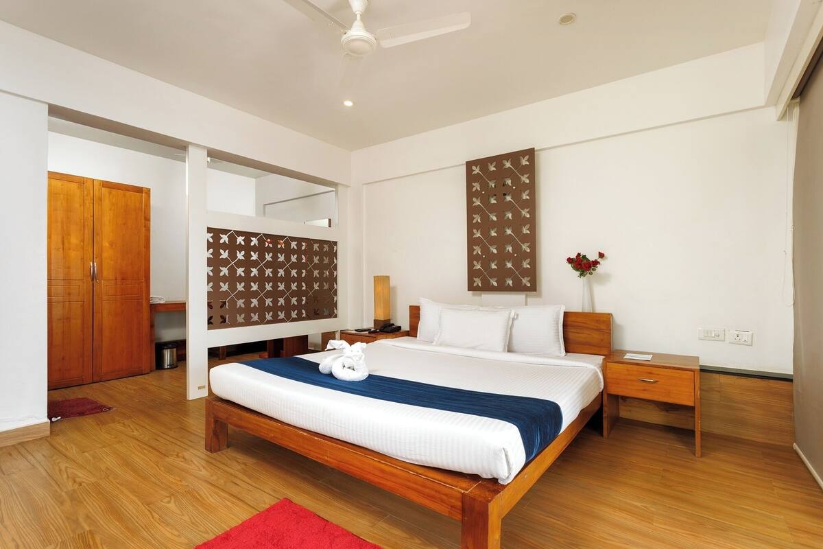 A modern hotel room with a wooden bed, a red rug, a wooden wardrobe, a nightstand, a wall-mounted decorative panel, and a ceiling fan.