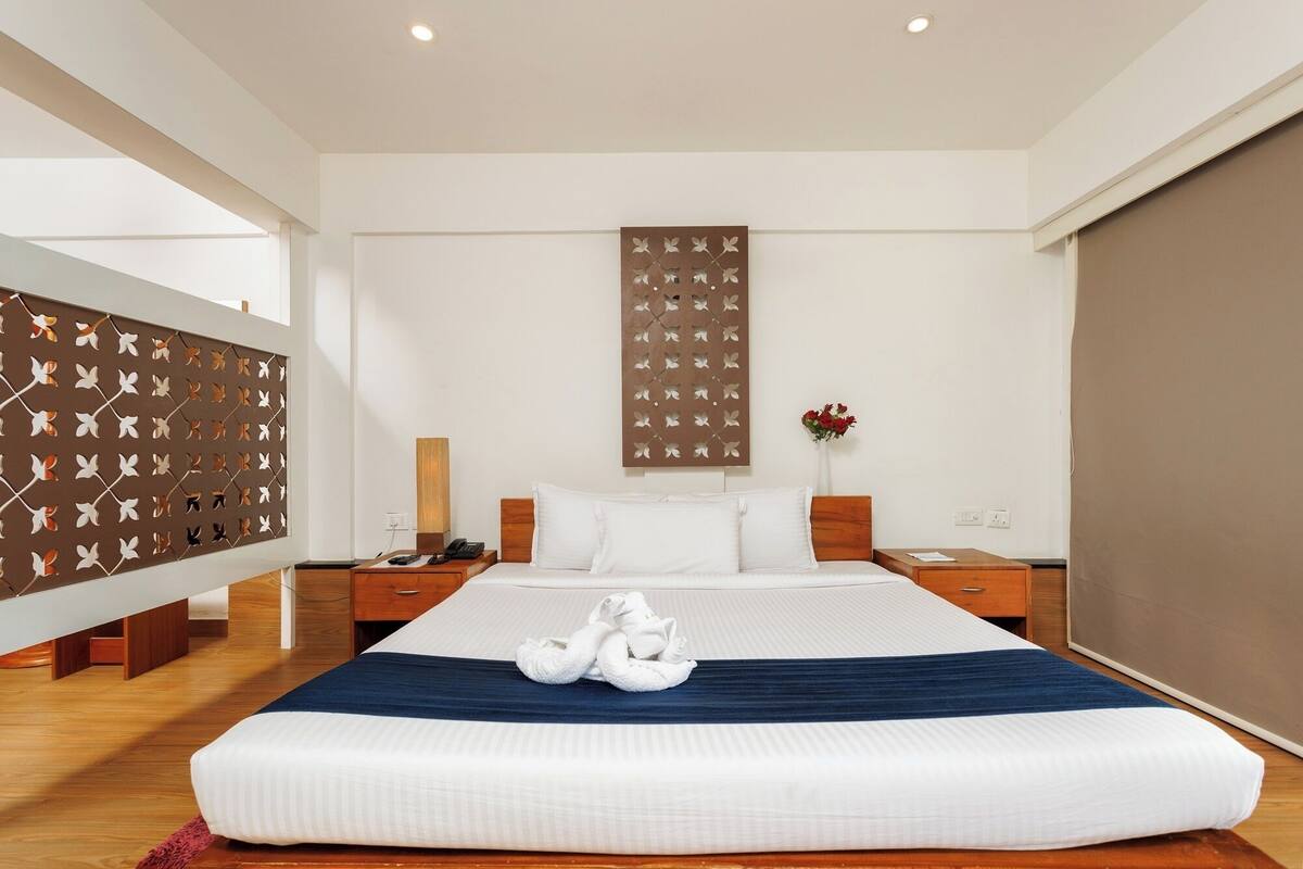 A modern hotel room with a large bed, wooden headboard, a decorative wall panel, a bedside table with a lamp, and a large window.