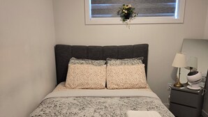A bedroom with a bed, a nightstand, a lamp, and a window with blinds. - Parkside Basement Suite/Netflix, Amazon Prime & Disney  (Edmonton)