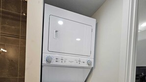 A compact laundry room with a front-loading washer and dryer, a tiled backsplash, and a door leading to a kitchen area. - Parkside Basement Suite/Netflix, Amazon Prime & Disney  (Edmonton)