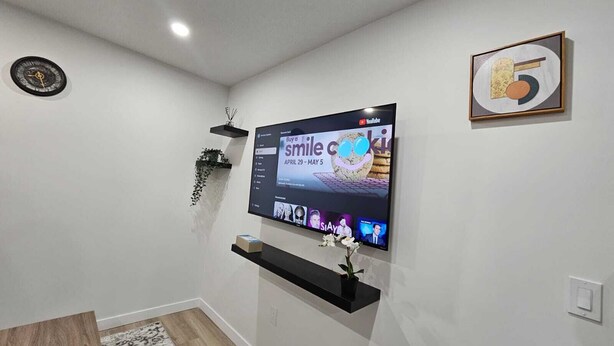 A modern living room with a flat-screen TV mounted on the wall, a black shelf below it, and a small plant on the shelf. - Parkside Basement Suite/Netflix, Amazon Prime & Disney  (Edmonton)