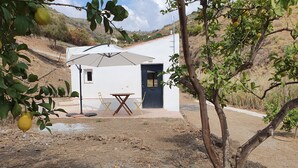 Um pequeno edifício branco com porta e janelas, uma mesa com cadeiras e uma árvore com limões.