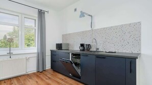 Cozinha moderna com armários cinzas, bancada branca, máquina de lavar louça embutida, chaleira e janela com cortinas.