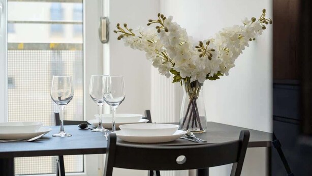 A dining table with white plates, two wine glasses, and a vase with white flowers.