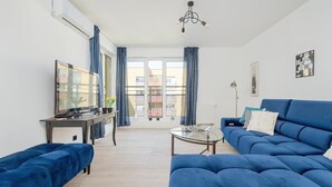 A living room with a blue sofa, a glass coffee table, a TV on a stand, and a balcony with curtains.