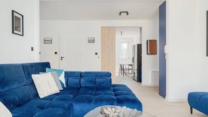 A modern living room with a blue sofa, a glass coffee table, and a dining area in the background.