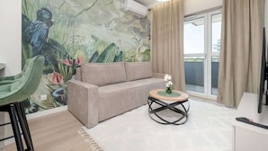 Ein Wohnzimmer mit einem beigen Sofa, einem grünen Sessel, einem runden Couchtisch mit einer Blumenvase und einem Wandbild von Vögeln und Pflanzen.