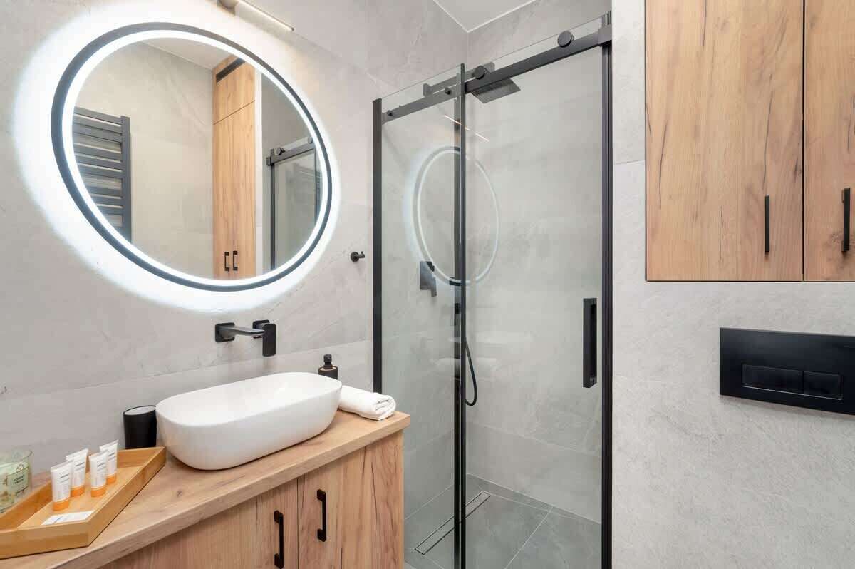 A modern bathroom with a round mirror, a glass shower enclosure, and wooden cabinetry.