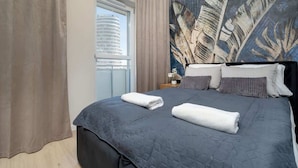 Ett hotellrum med en stor säng, utsikt över en byggnad genom ett fönster och en vägg prydd med ett tropiskt bladmotiv.