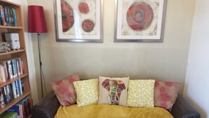 Un salon avec un canapé en cuir noir, un plaid jaune et des coussins colorés. On y trouve une bibliothèque remplie de livres et un lampadaire rouge. Deux tableaux abstraits sont accrochés au mur, au-dessus du canapé.