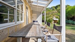 An outdoor covered patio with a wooden table and chairs, surrounded by a garden.