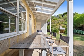 An outdoor covered patio with a wooden table and chairs, surrounded by a garden.