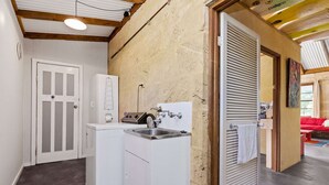 A compact laundry area with a washing machine, a sink, and a door leading to a living space. - Kangaroo Cottage – Beachside Escape in Dawesville (Dawesville)