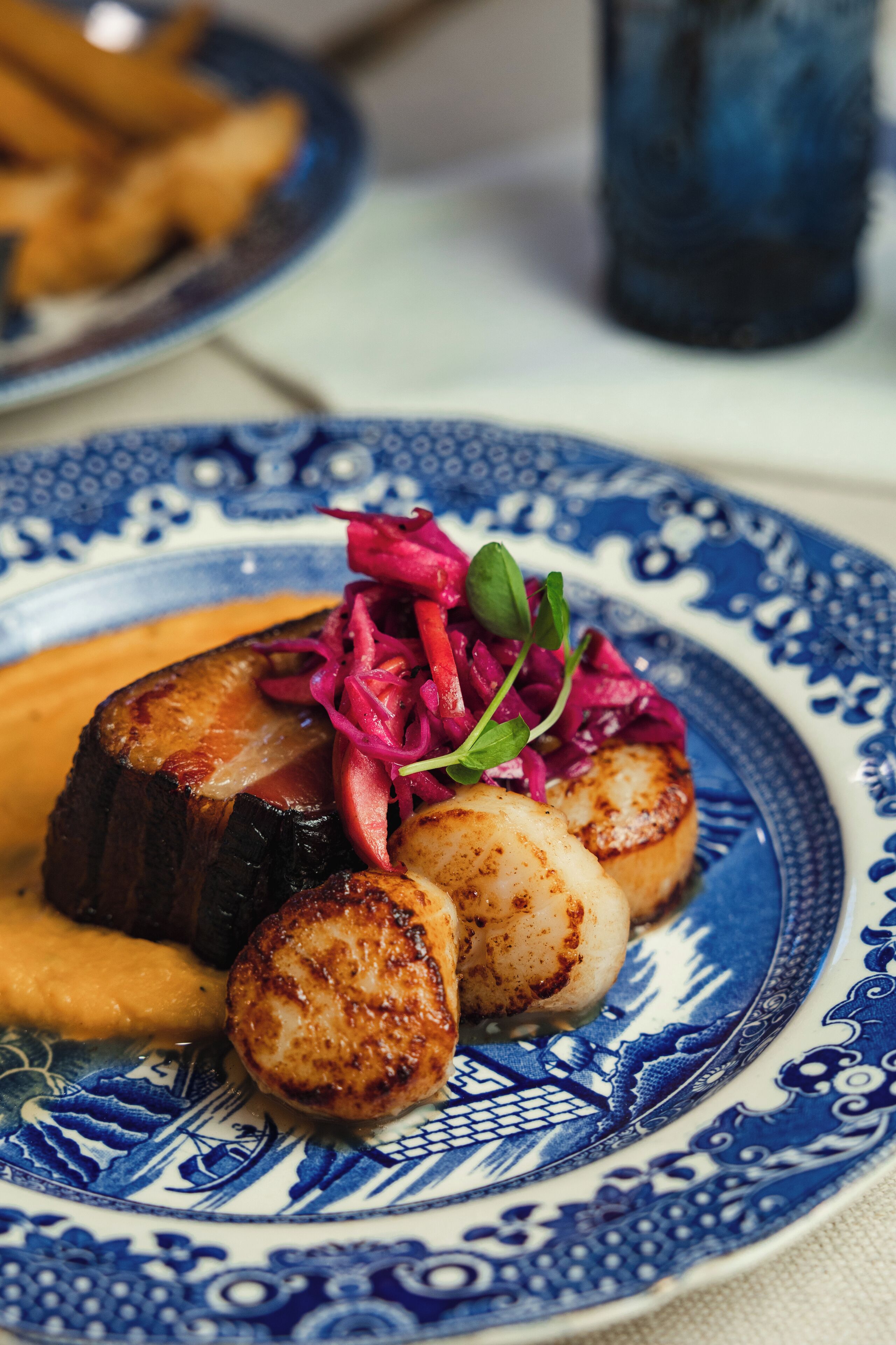 A plate with seared scallops, roasted potatoes, and a beetroot salad on a blue and white patterned plate.