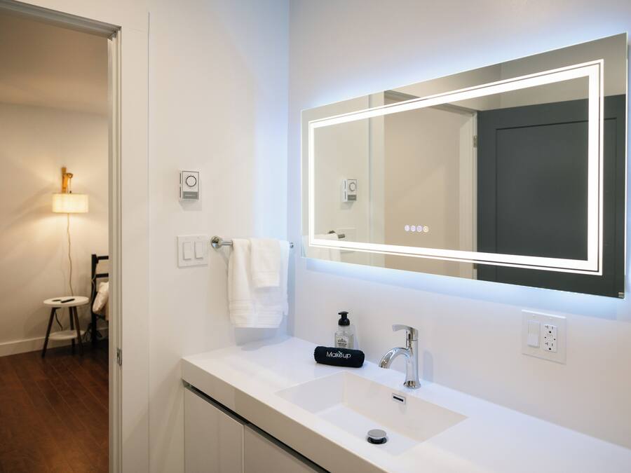 A modern bathroom with a white sink, a large mirror with integrated lighting, a handheld soap dispenser, and a towel hanging on a rack.