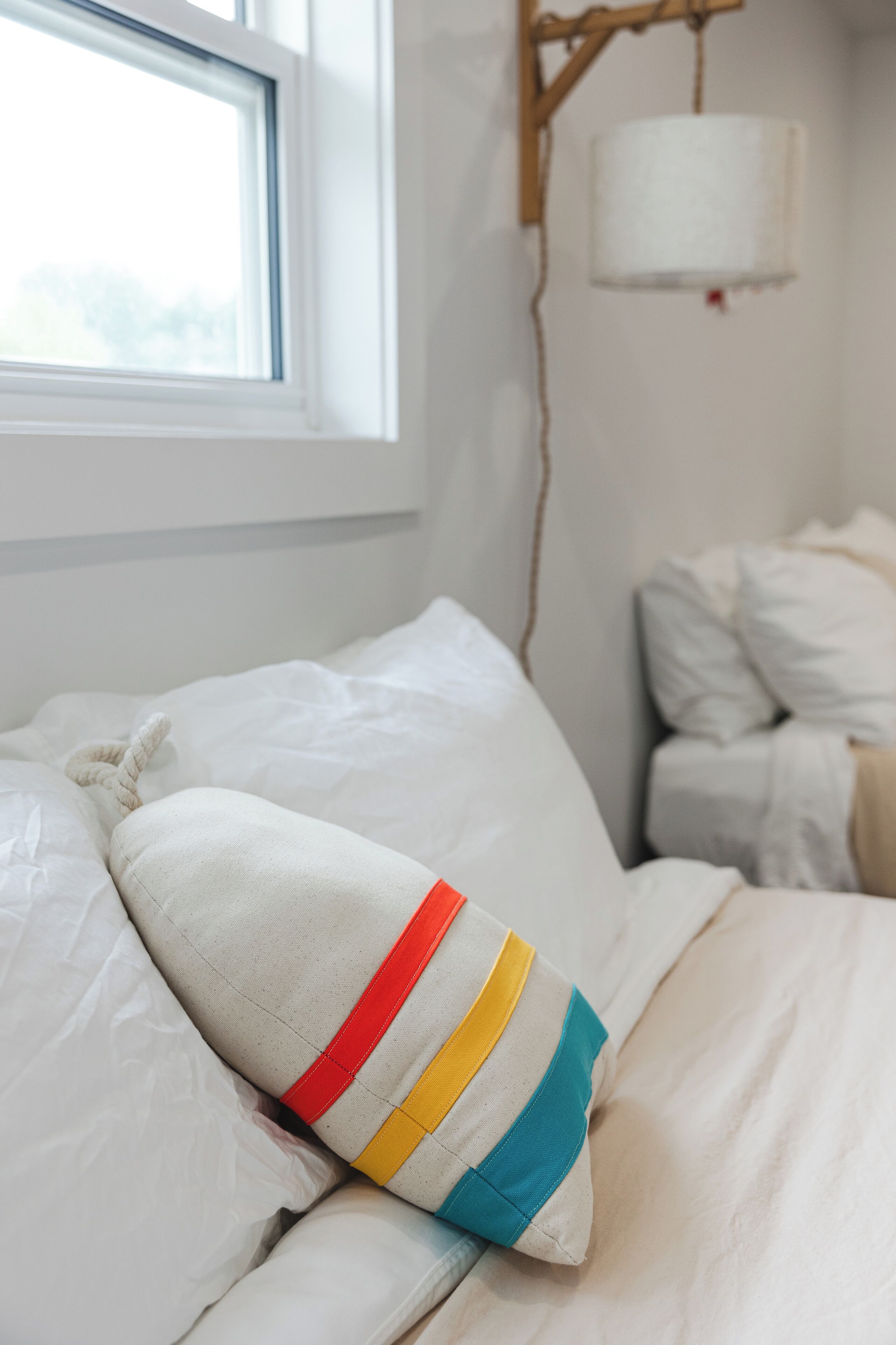 Una cama con ropa blanca, una almohada colorida y una ventana con una lámpara blanca.