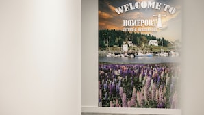 A welcome sign for a hotel with a scenic view of a lake and boats, and a field of purple flowers below. - Homeport Suites & Residences (Lords Cove)