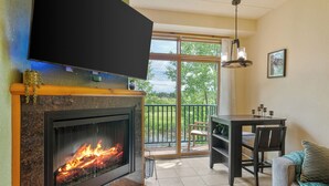 A living room with a fireplace, a TV mounted on the wall, a dining table with chairs, and a balcony with a view of greenery. - Tee Time Terrace - Near Attractions / Waterpark (Wisconsin Dells)