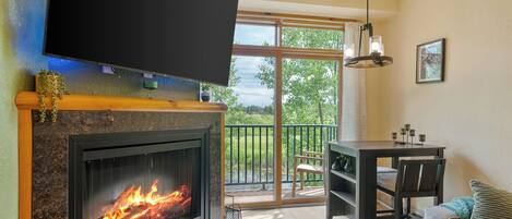 A living room with a fireplace, a television mounted above it, a dining area with a table and chairs, and a balcony with a view of greenery.