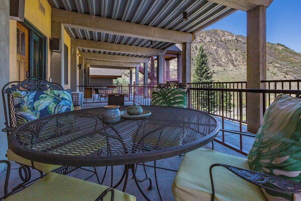 A covered outdoor seating area with a round table and chairs, a view of a mountainous landscape, and a building with a brick facade in the background.
