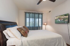 A bedroom with a bed, a ceiling fan, a window with curtains, a TV on the wall, and a lamp.