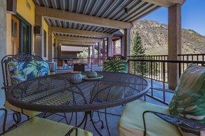 A covered outdoor seating area with a round table and chairs, a view of a mountainous landscape, and a building with a brick facade in the background.
