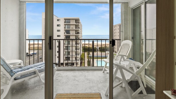 Un balcon avec des chaises en plastique blanches, une petite table et une vue sur l’océan et les bâtiments.