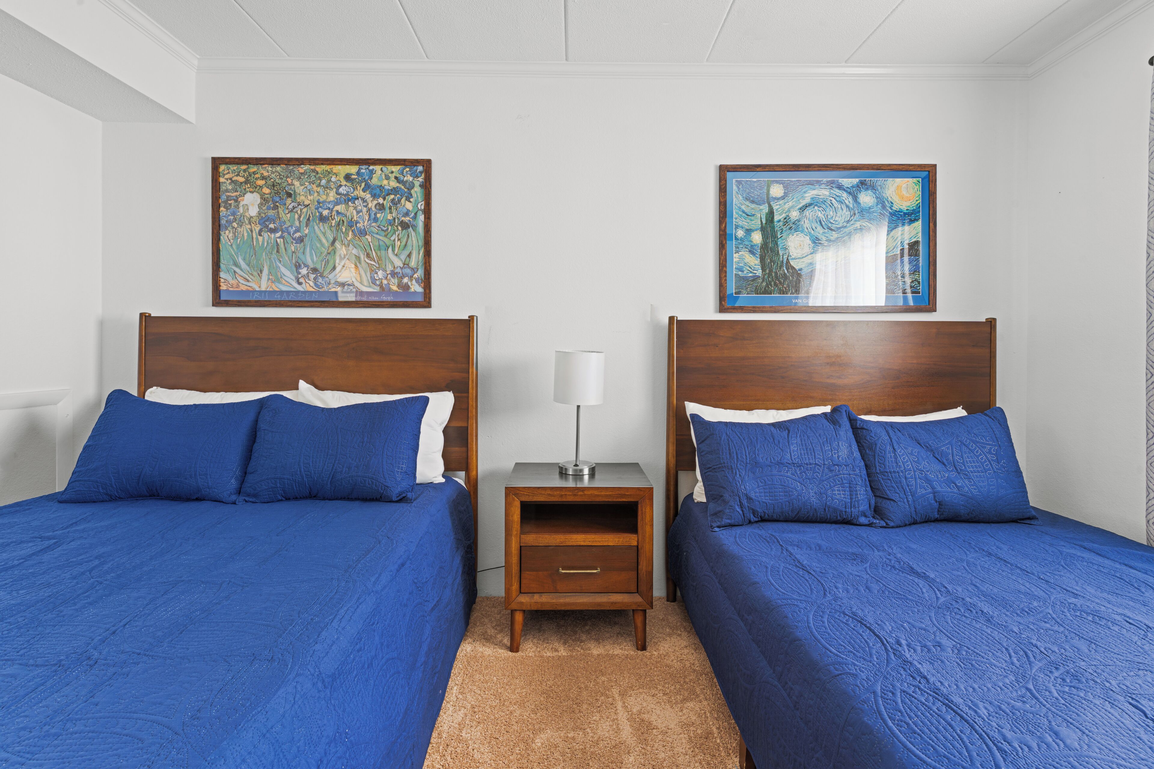 A room with two beds, each with a wooden headboard and blue bedding. There is a nightstand between the beds with a lamp.